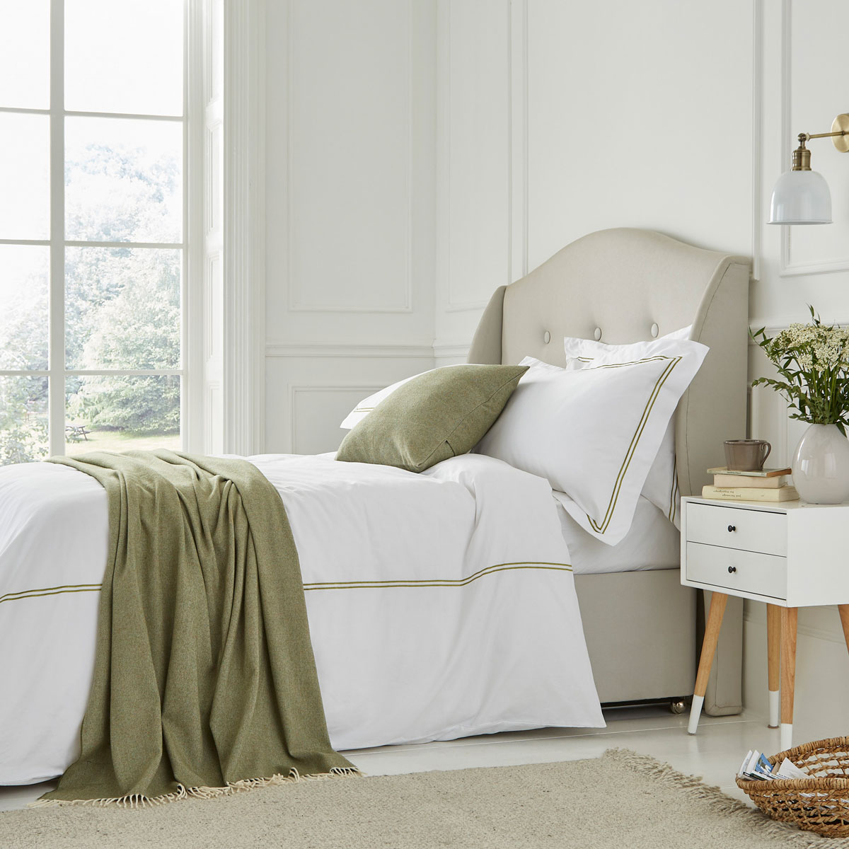 Spálňa so smotanovou posteľou s vysokým čalúneným čelom a bielymi obliečkami s olivovozeleným orámovaním