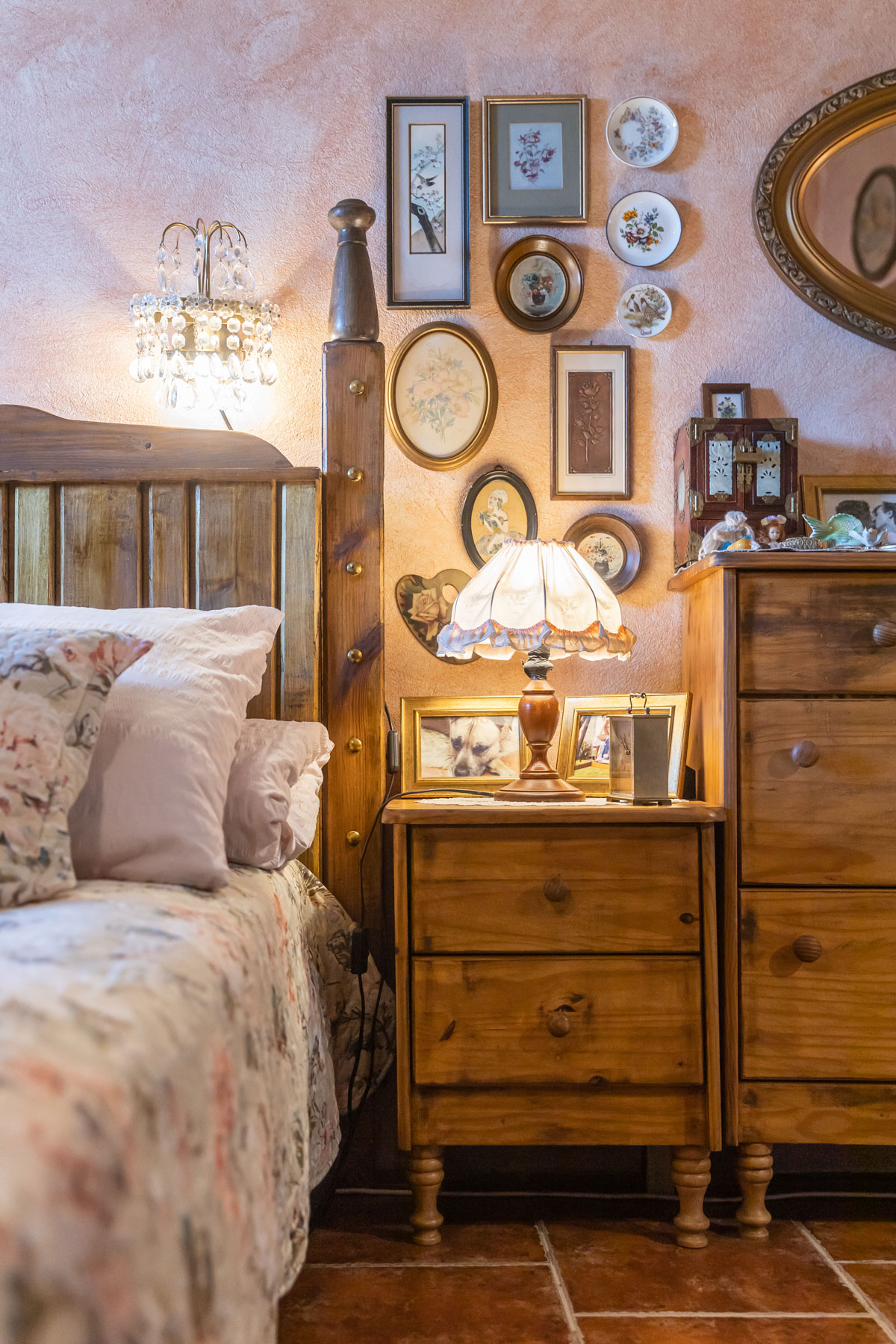 detail spálne vo vidieckom štýle, na obrázku vidieť drevený nočný stolík so starožitnou lampou, po jednej strane stolíka stojí väčšia zásuvková skrinka, po druhej strane je drevená posteľ s ozdobným okrajom čela, na stene visia staré obrazy a tanieriky