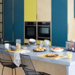Minimalistická farebná kuchyňa, kuchynská stena má dvierka z drevodekóru, niektoré časti sú modré a jasnožlté, oproti je ostrovček s čiernymi stoličkami
