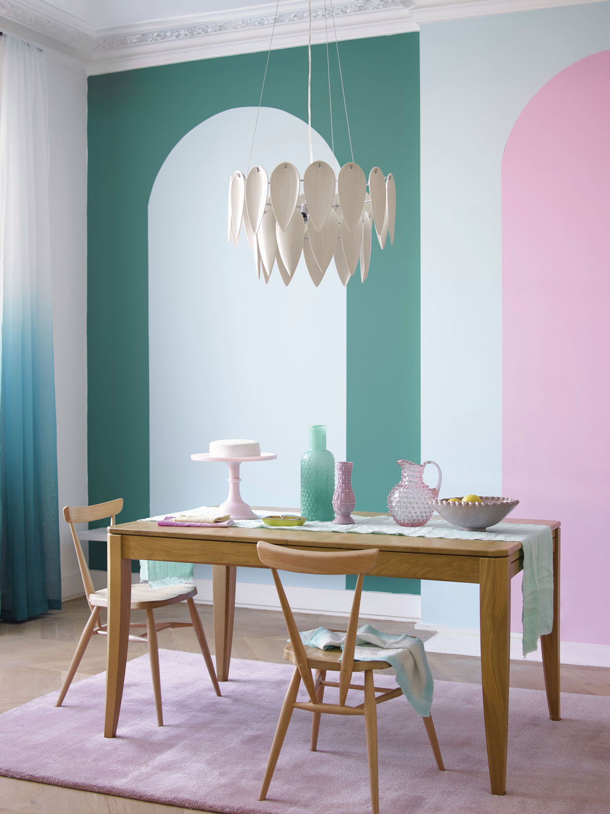 Jedáleň s farebnými fialovo-zelenými stenami, dizajnovým lustrom a jednoduchým dreveným stolom so stoličkami, na stole sú sklenené nádoby v zelenej a fialovej farbe