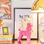 veselý detail z interiéru módnej návrhárky Mariny Hoermanseder, na polici vidieť extravagantnú lampu so zlatým tienidlom a nohou s farebnými sklenými bublinami, vedľa je dekorácia ružového pudlíka, knihy o umelcoch, extravagantná misa s drobnými hračkami a nad ňou visí abstraktný farebný obraz