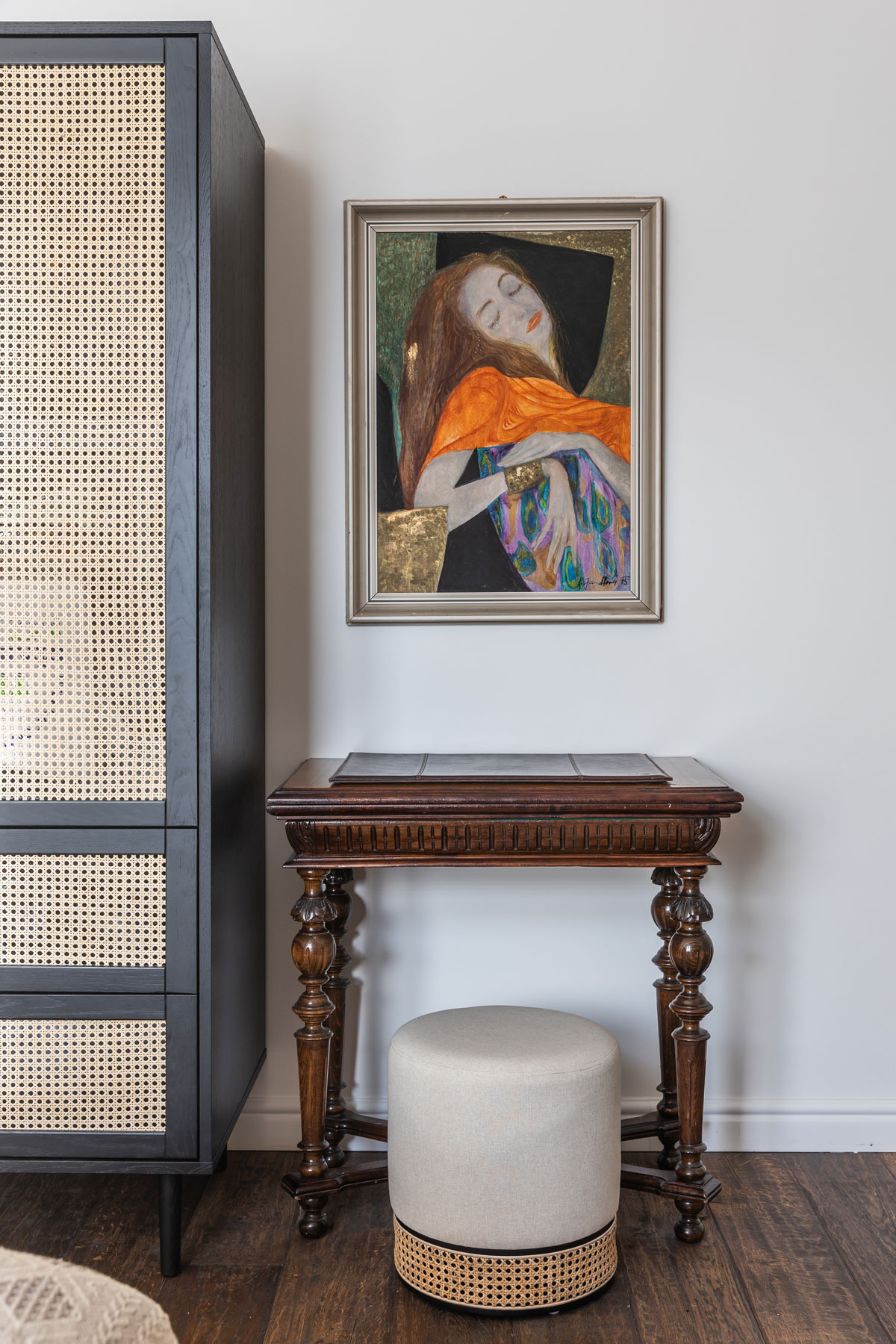 detail interiéru s čiernou skriňou s viedenským výpletom, vedľa nej stojí starožitný stolík s modernou svetlou taburetkou, nad stolíkom, je obraz ženy v secesnom štýle