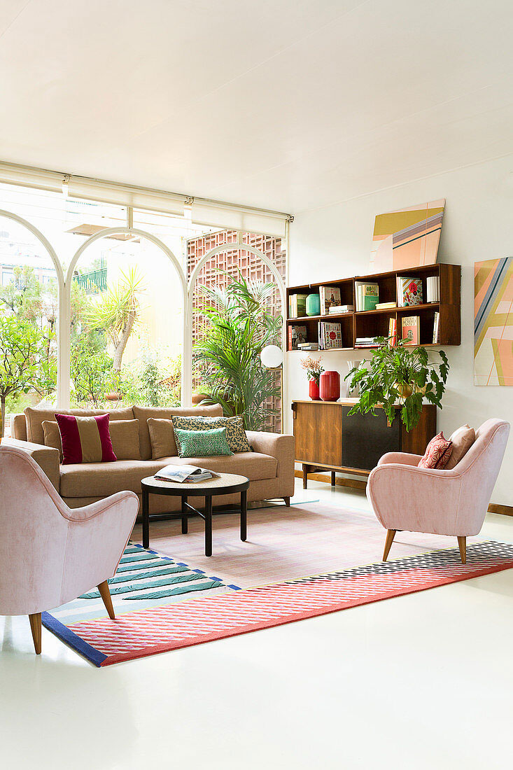 Obývačka v pastelových farbách, v strede je svetlohnedá sedačka s tmavým dreveným konferenčným stolíkom, oproti stoja dve zamatové ružové kresielka, tento nábytok spája farebný vzorovaný koberec, pri stene je retro komoda a otvorená skrinka s knihami