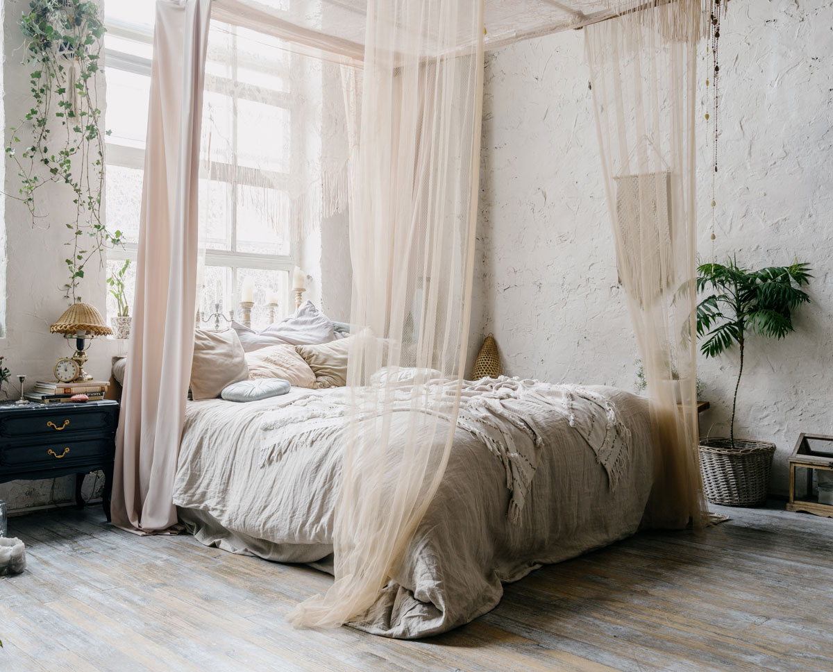 Spálňa v prírodnom štýle, posteľ je ustlaná v prírodných materiáloch a svetlých farbách, okolo postele sú závesy a záclony proti hmyzu, vedľa postele je tmavomodrý retro stolík