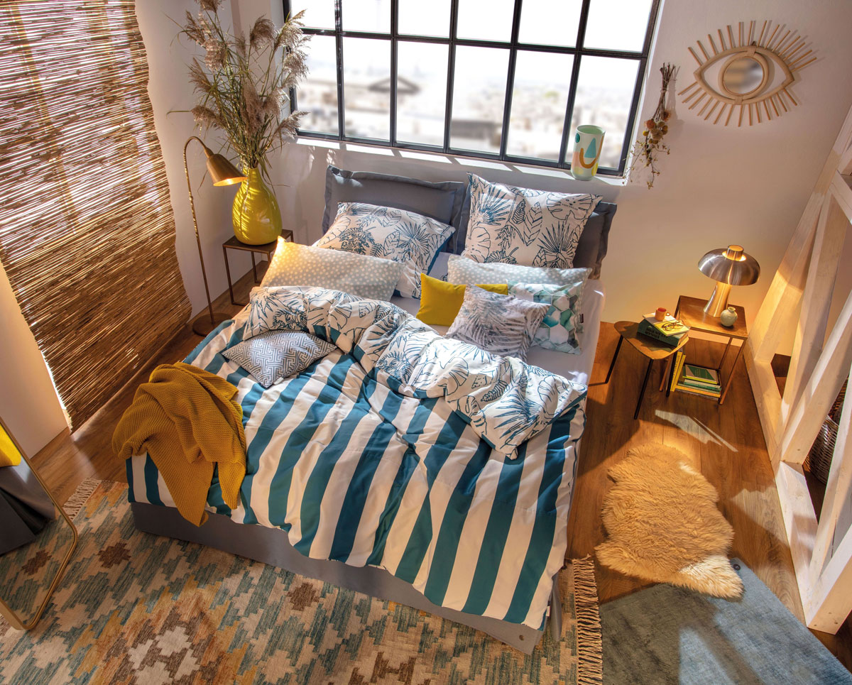 Spálňa so vzorovaným kobercom, čalúnenou posteľou a farebnými modro-bielymi obliečkami so vzorom rastlín z jednej strany a pásikmi z druhej strany