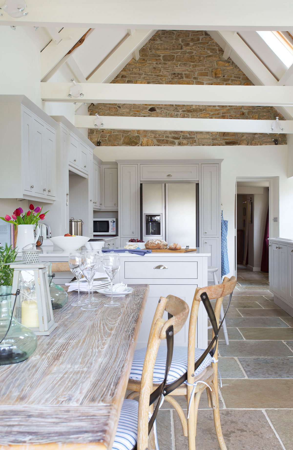 škandinávska vidiecka kuchyňa s bielou kuchynskou linkou do tvaru písmena U, pred polostrovom je drevený stôl s drevenými stoličkami, na strope sú priznané trámy a kameň