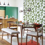 retro moderná jedáleň, na jednej stene je retro zeleno-biela tapeta, v strede je elegantný dizajnový stôl s modernou verziou retro stoličiek, stôl stojí na svetlom koberci s farebnými kruhmi, za jedálňou vidieť kuchyňu s ostrovom a zelenú skriňu