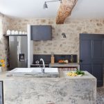 kuchyňa v chalupe s priznanou kamennou stenou a jednoduchou modernejšou kuchynskou linkou, v ktorej dominujú povrchy z betónu vrátane ostrovčeka, dvere do špajze sú antracitové, chladnička je od sporáku oddelená stenou
