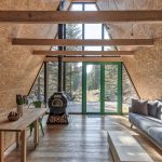 pohľad do interiéru typickej horskej chaty s áčkovou strechou, ktorá je z jednej časti presklená s výhľadom do lesa a vstupom na terasu, v interiéri je jednoduchá sivá sedačka, minimalistický stolík a piecka