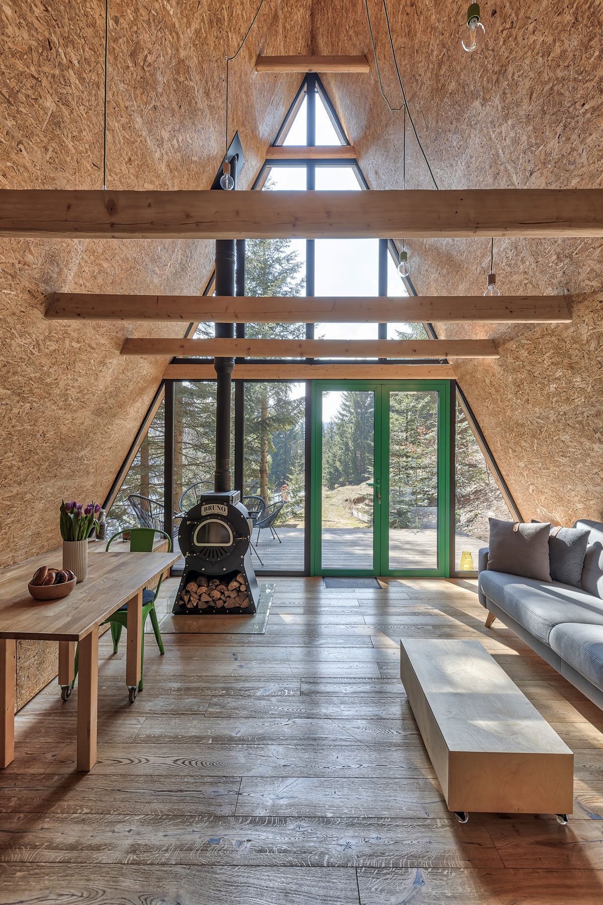 pohľad do interiéru typickej horskej chaty s áčkovou strechou, ktorá je z jednej časti presklená s výhľadom do lesa a vstupom na terasu, v interiéri je jednoduchá sivá sedačka, minimalistický stolík a piecka