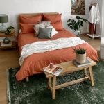 spálňa v etno štýle s drevenou posteľou, na ktorej sú obliečky v broskyňovej farbe, pred posteľou je drevená lavička, na jednej strane postele je drevený nočný stolík, na podlahe je tmavozelený orientálny koberec