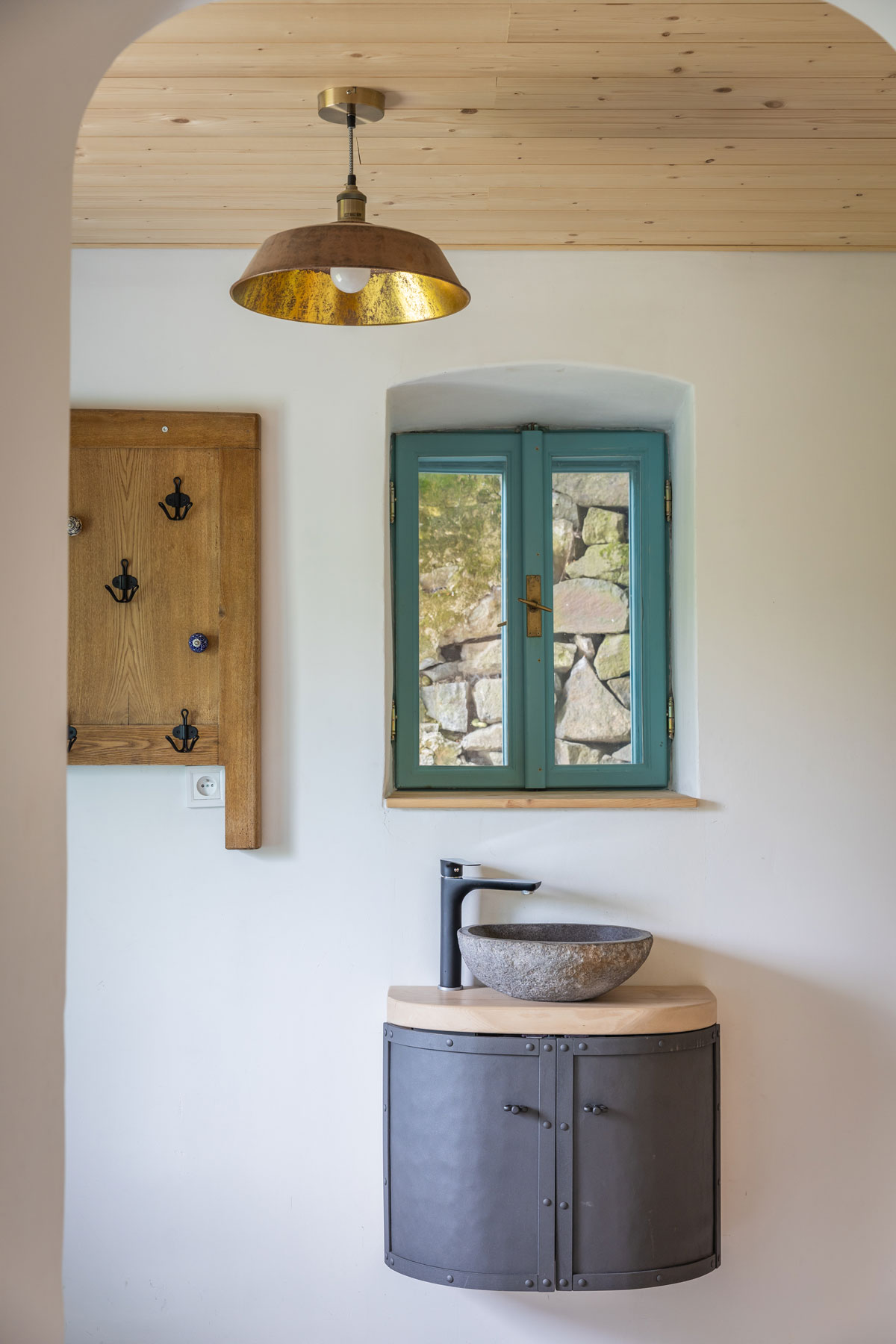 jednoduchá kúpeľňa v chalupe s oknom v rámom natretým na modro, pod oknom je zrecyklovaná kovová skrinka s malým umývadlom, vedľa okna visí vešiak vyrobený z rámu postele