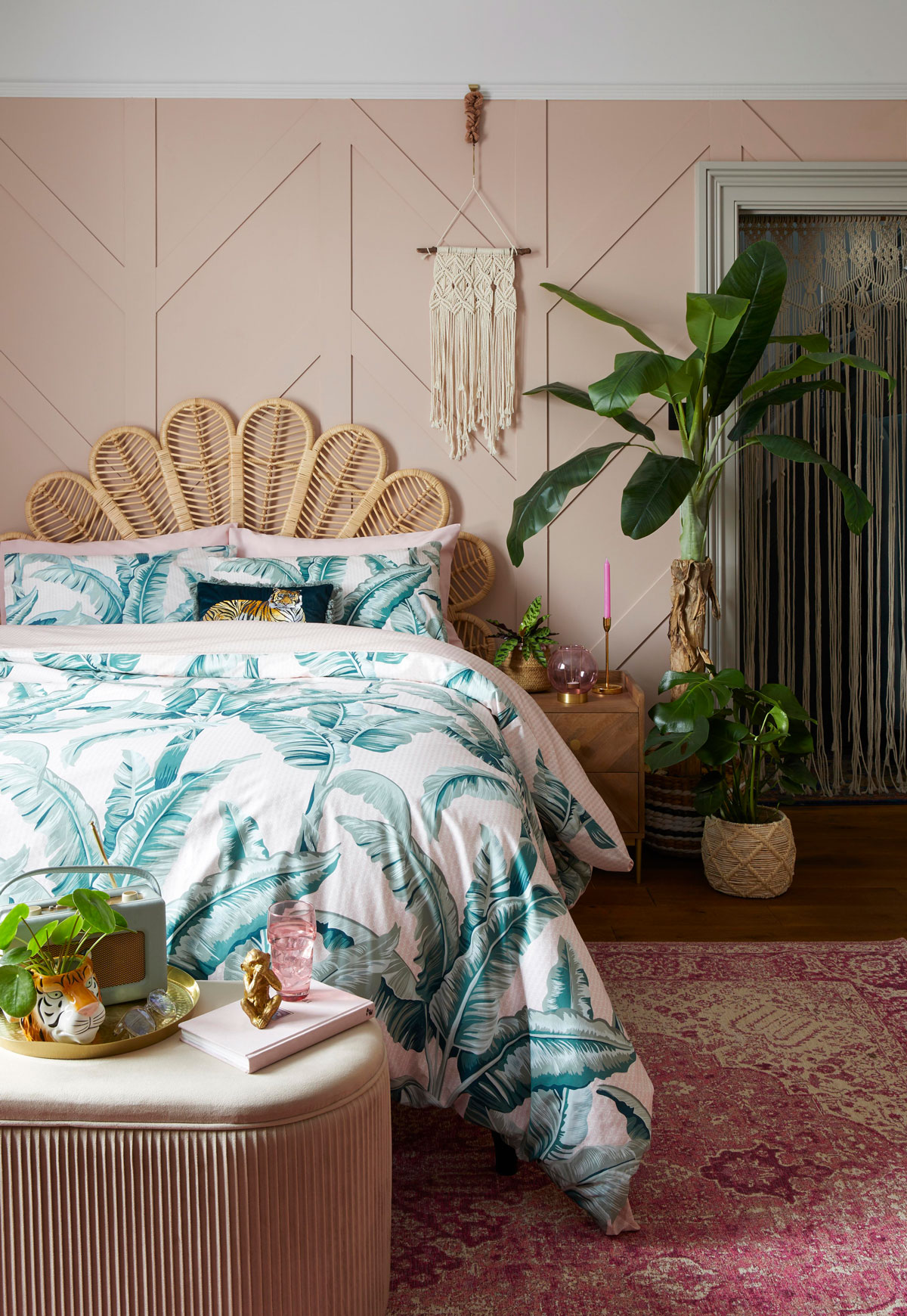 Spálňa s izbovými rastlinami zariadená v tropickom štýle, na zemi je tkaný orientálny koberec, posteľ má čelo v tvare listu a je ustlaná s obliečkami s motívom exotických rastlín, vedľa je drevený nočný stolík, štýl dopĺňajú ozdoby so zvieratami z džungle