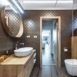 elegantná hnedá kúpeľňa so štruktúrovanými obkladmi na stenách, v kúpeľni je na jednej stene závesná drevená skrinka s umývadlom, oproti je závesné WC, nad umývadlom je veľké okrúhle zrkadlo