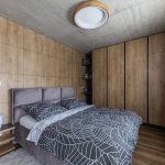 elegantná moderná spálňa so sivou zamatovou posteľou, vstavané skrine sú z dreva, na stenách je drevený obklad, výrazným prvkom je betónový strop