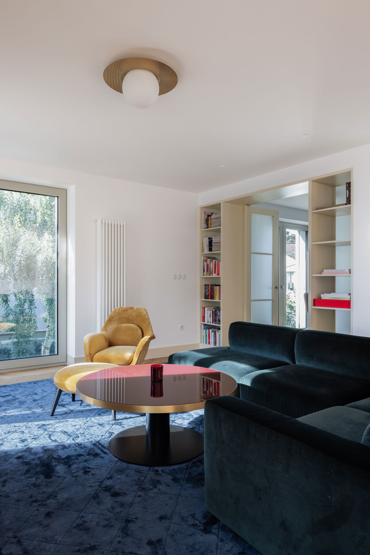 obývačka v klasickom glamour štýle, dominuje jej celoplošný tmavomodrý koberec, na ňom stojí výrazný konferenčný stolík, zamatové žlté kreslo a zamatová sedačka v kráľovskej modrej, z obývačky vedú dvojkrídlové dvere