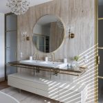 elegantná glamour kúpeľňa s travertínovým obkladom, na stene je veľké okrúhle zrkadlo v jemnom zlatom orámovaní, po oboch jeho stranách sú dve lampy, umývadla s mosadznými batériami sú posadené do kamennej dosky, pod ňou je biela skrinka na nožičkách