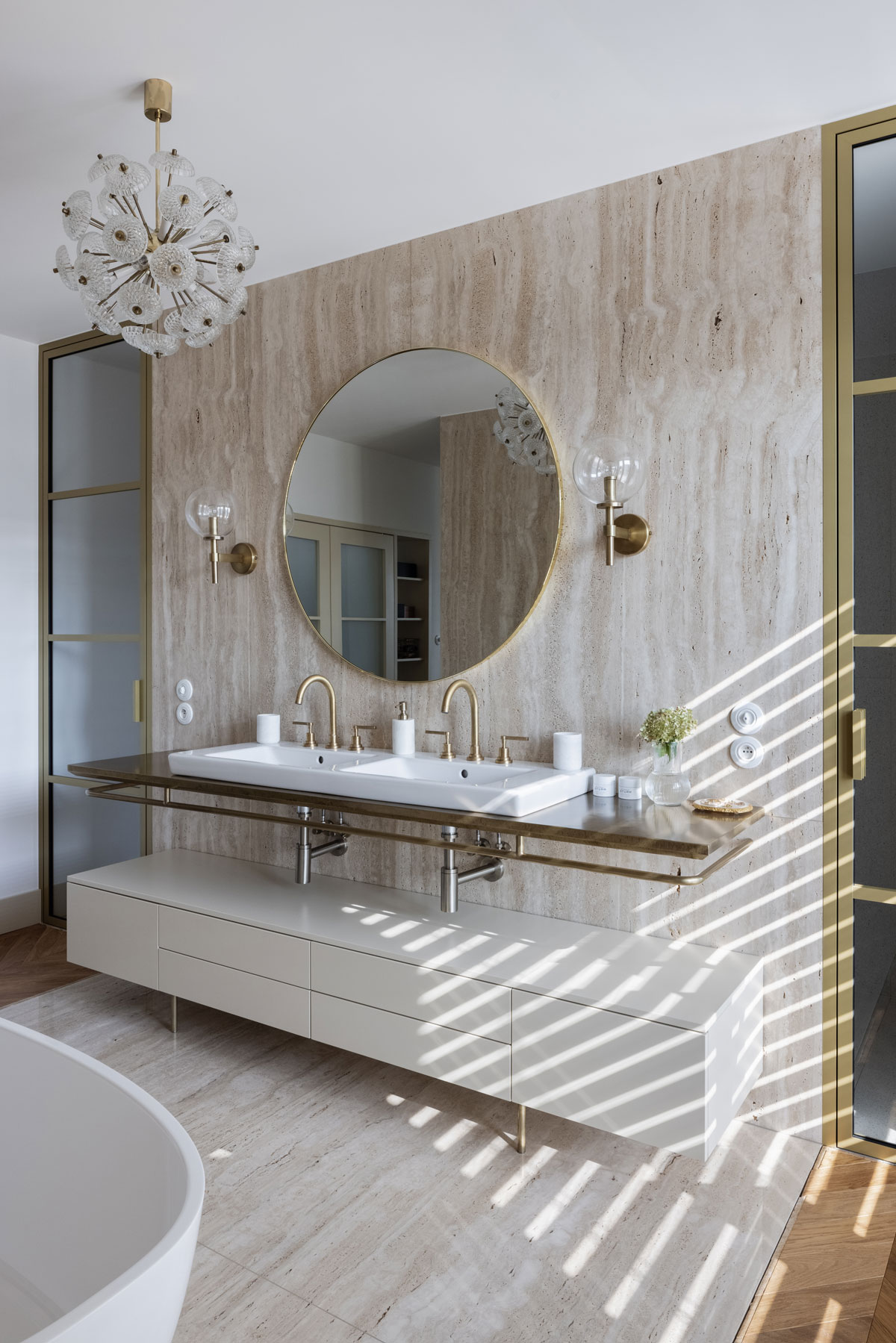 elegantná glamour kúpeľňa s travertínovým obkladom, na stene je veľké okrúhle zrkadlo v jemnom zlatom orámovaní, po oboch jeho stranách sú dve lampy, umývadla s mosadznými batériami sú posadené do kamennej dosky, pod ňou je biela skrinka na nožičkách