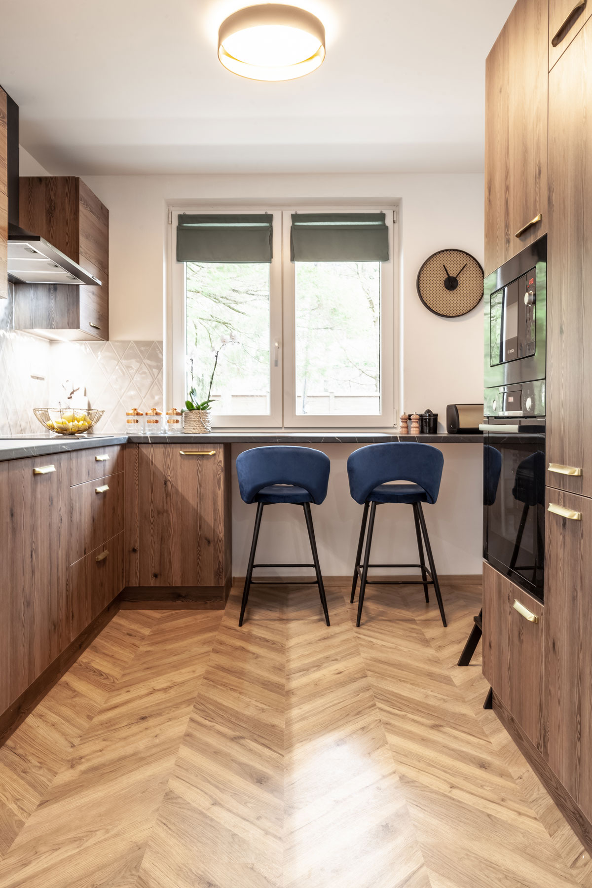 klasická moderná kuchyňa v tmavom drevodekore, nábytok v kuchyni má zlaté úchytky, pri pracovnej doske pod oknom sú dve barové stoličky v modrom zamate