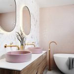 Moderná kúpeľňa s ružovým obkladom na stene a sivou dlažbou, na stene visí drevená skrinka s ružovými umývadlami, zo steny sú vyvedené zlaté batérie