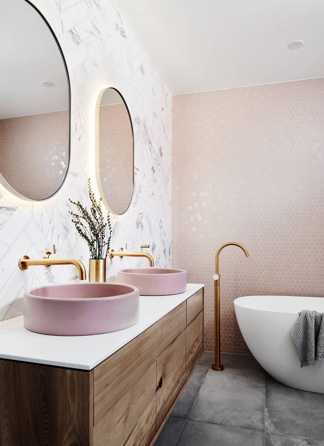Moderná kúpeľňa s ružovým obkladom na stene a sivou dlažbou, na stene visí drevená skrinka s ružovými umývadlami, zo steny sú vyvedené zlaté batérie