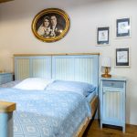 zrenovovaná stará posteľ s modrým otapetovaným čelom, vedľa nej stojí nočný starodávny stolík v rovnakom prevedení ako čelo postele nad posteľou je starý obraz v oválnom ráme