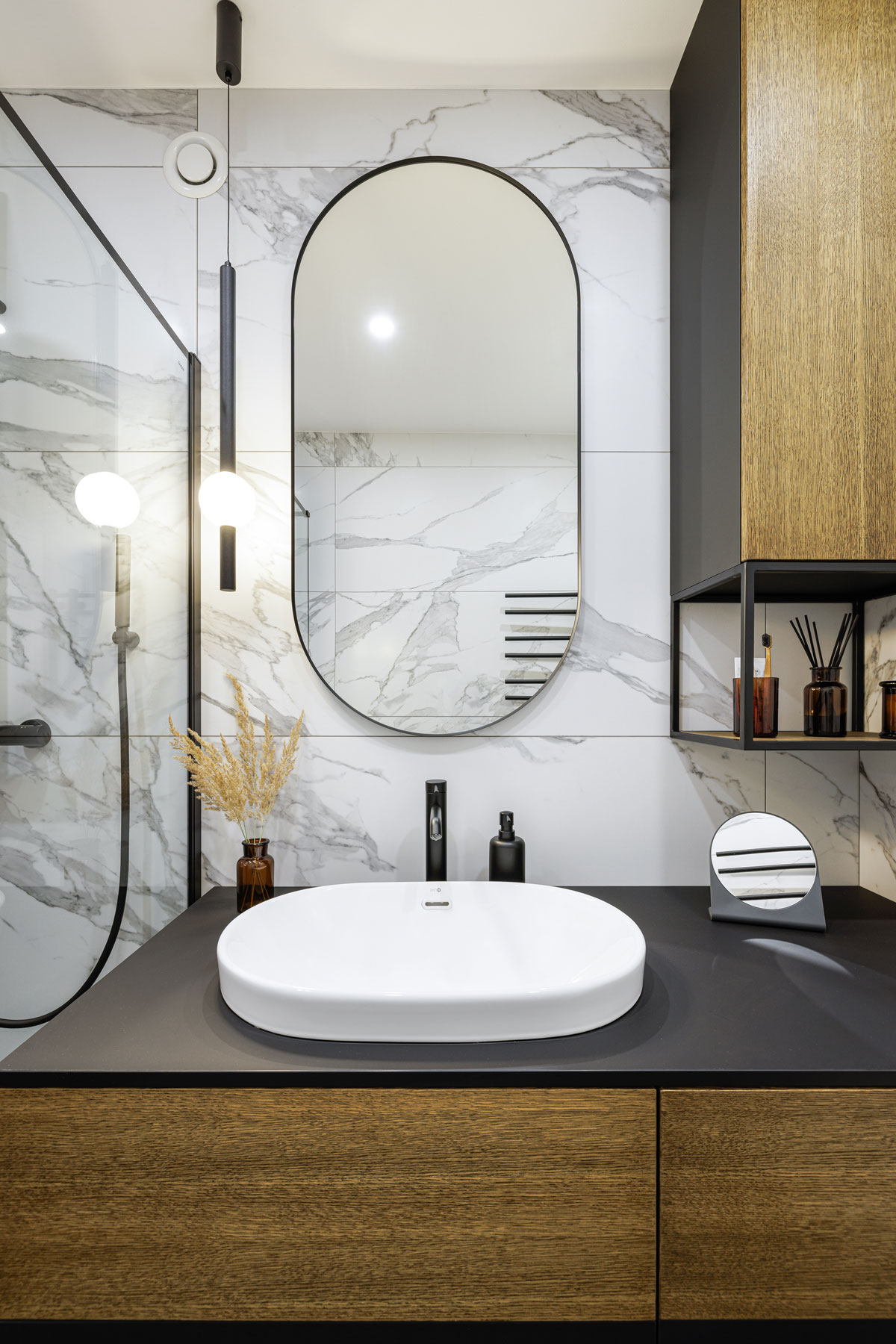 moderná kúpeľňa s mramorovým obkladom, skrinkou s umývadlom, ktorá má čiernu dosku a dvierka z drevodekóru, na tene visí oválne zrkadlo a kovová skrinka v rovnakom ladení ako umývadlová
