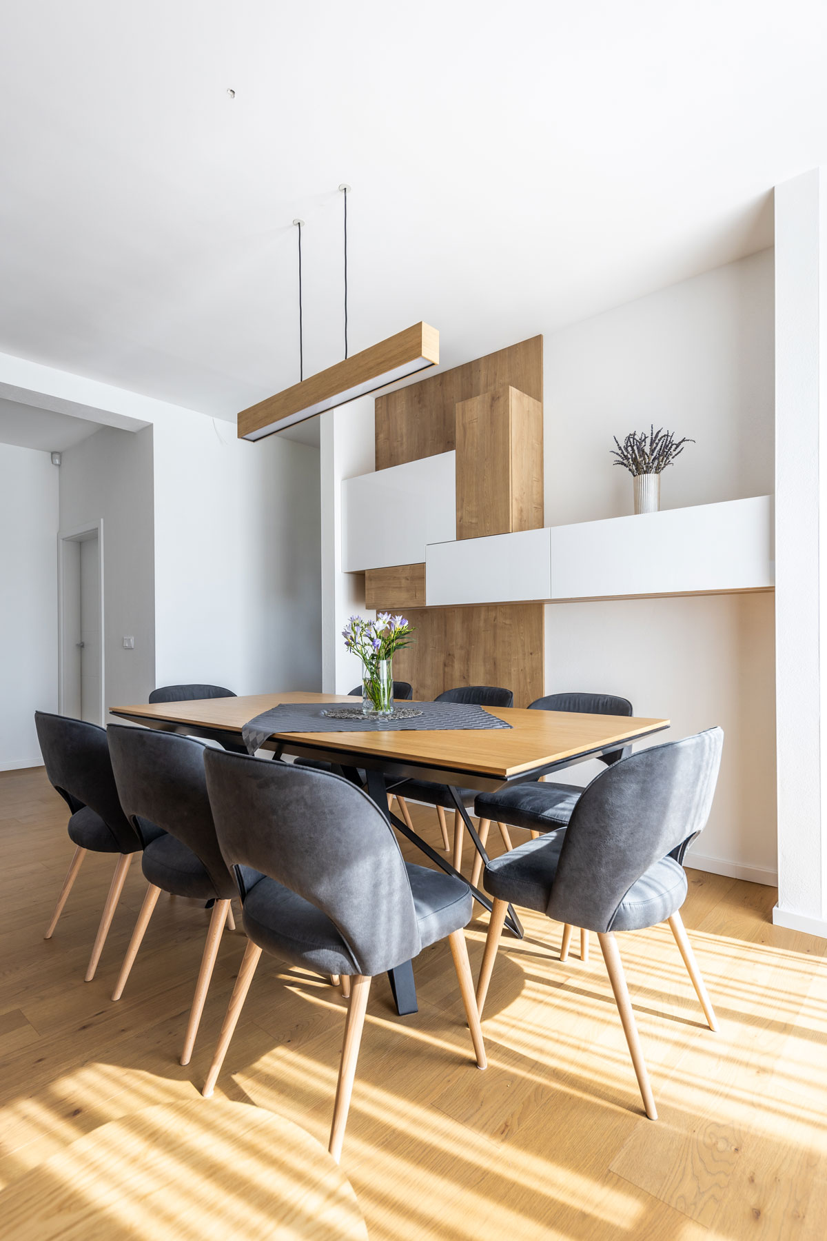 jedáleň v rodinnom dome s dreveným stolom a sivými čalúnenými stoličkami, za stolom je stena s obdĺžnikovými skrinkami v akoby kubistickom rozložení