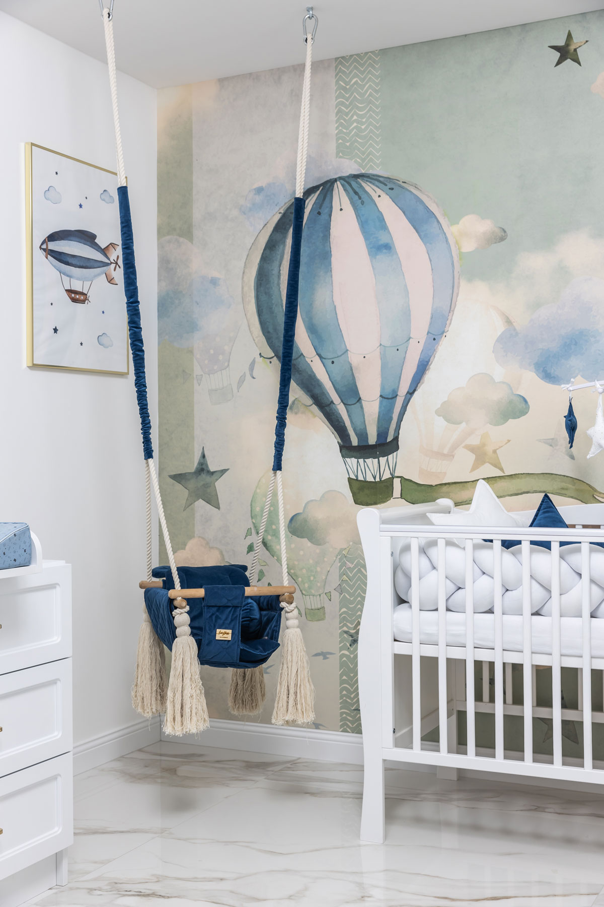 časť detskej izby ladená do modro-bielej kombinácie s bielou postieľkou, modrou hojdačkou a tapetou s motívom teplovzdušných balónov