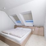 minimalistická podkrovná spálňa s drevenou posteľou, skrinkami pod šikminou a na mieru vyrobenými vstavanými skriňami, stena naproti skriniam je tehlová natretá nabielo