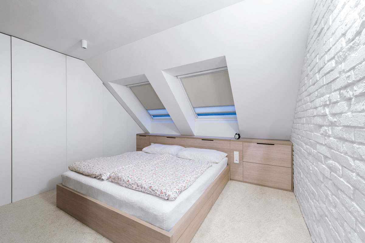 minimalistická podkrovná spálňa s drevenou posteľou, skrinkami pod šikminou a na mieru vyrobenými vstavanými skriňami, stena naproti skriniam je tehlová natretá nabielo