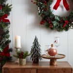 Sviatočný aranžmán na komode s malým vianočným stromčekom, umelými vetvičkami ihličia, podnosom s likérom a pohármi so zimným motívom, nad komodou visí ešte veniec