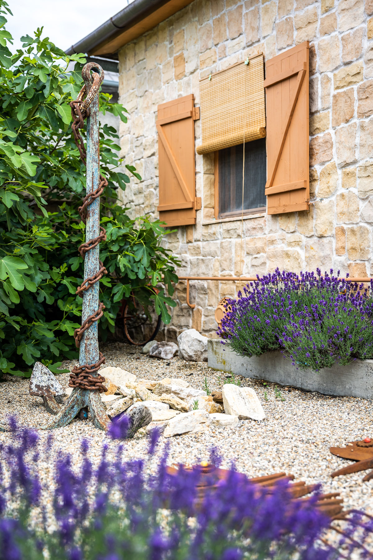 Časť záhrady vysypaná štrkom, v ktorom je zapichnutá obrovská kotva z lode, popri múre domu sa tiahne betónový kvetináč s levaduľou