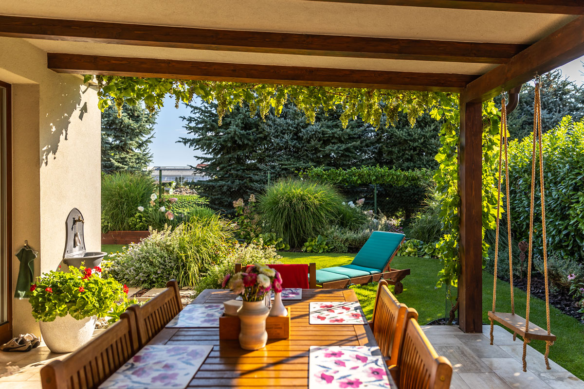 terasa v rodinnom dome s dreveným jedálenským stolom, na tráme zastrešenia terasy visí hojdačka, v pozadí vidieť záhradu s bujnou zeleňou a lehátko