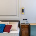 detail spálne s jednoduchou elegantnou posteľou, vedľa nej stojí zrenovovaný retro nočný stolík a farebná komoda, na stene sú ozdobné lišty