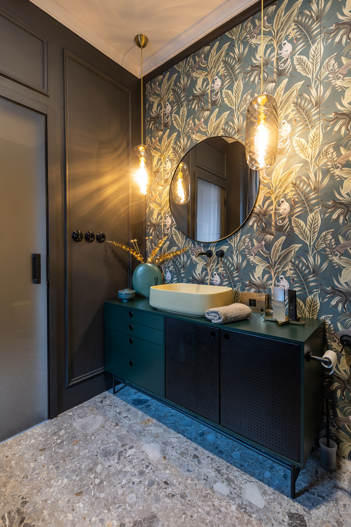 tmavá elegantná kúpeľňa s čiernou skrinkou a umývadlom v žltej farbe, stenu tvorí obklad s tropickým motívom s opicami, na stene je tiež zavesené okrúhle zrkadlo, kúpeľňu osvetľujú retro svietidlá, podlaha je imitáciou terazzových mramorových dlažieb