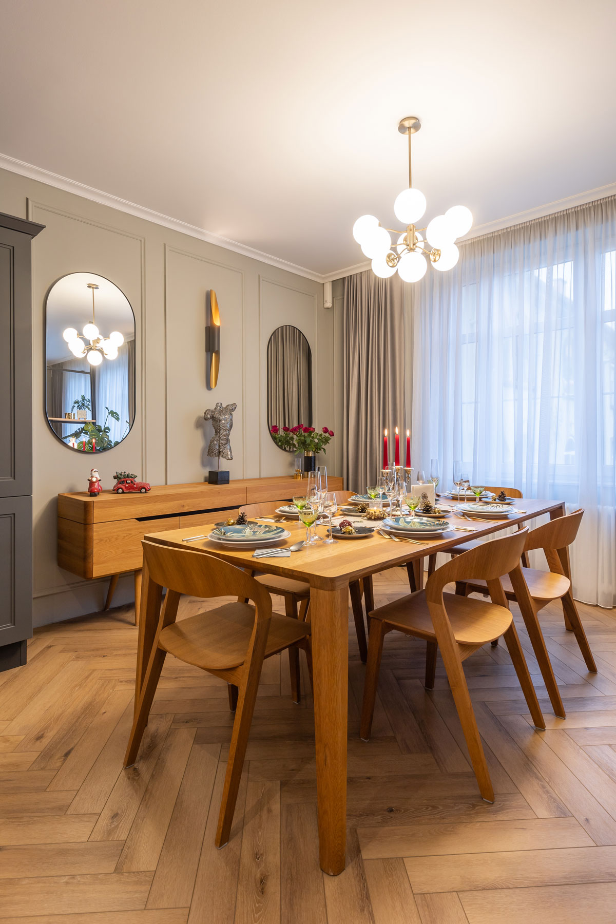 jedálenský stôl s retro štýle s dizajnovými stoličkami, prestretý sviatočne, nad stolom svieti zlatý retro luster, za stolom je pri stene retro komoda, steny zdobia lišty a oválne zrkadlá