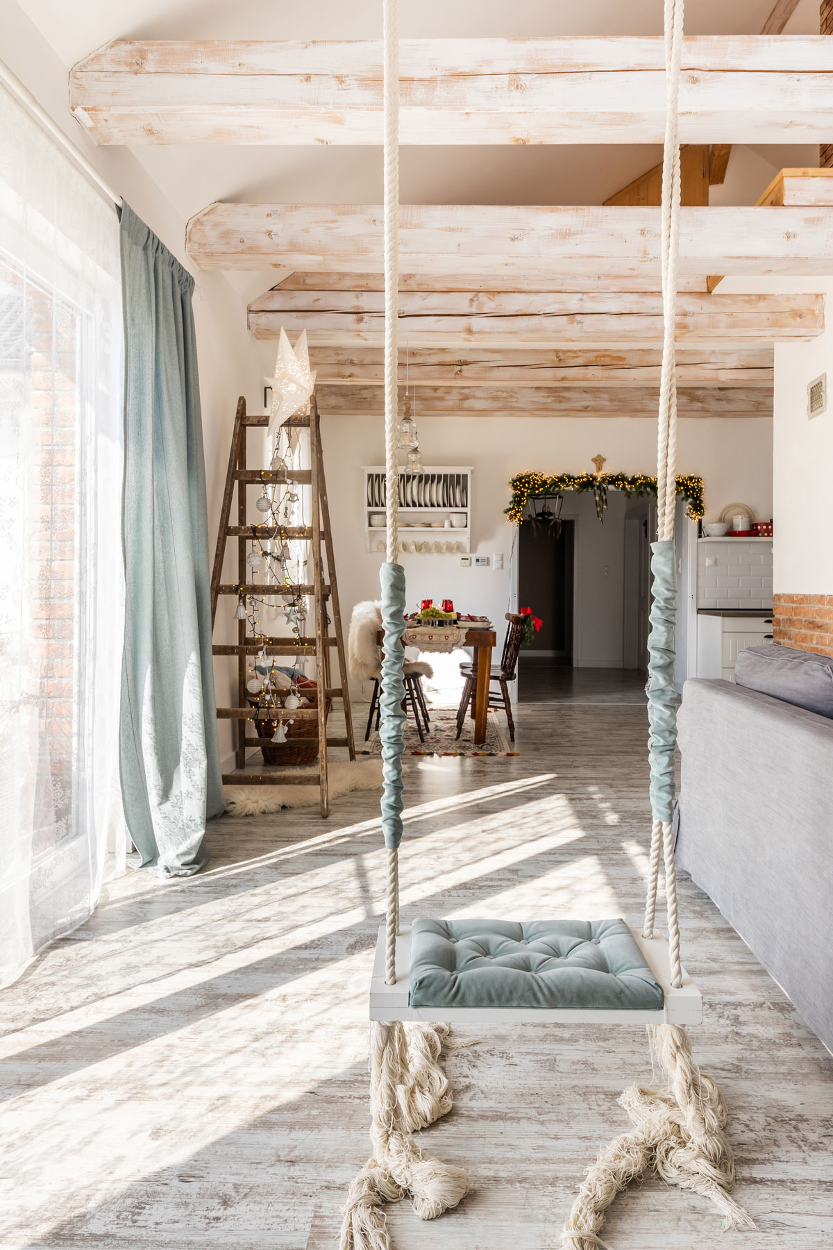 interiér v rodinnom dome s boho hojdačkou, ktorá visí z drevených trámov, v pozadí vidieť áčkový rebrík, na ktorom je vianočná hviezda a svetielka
