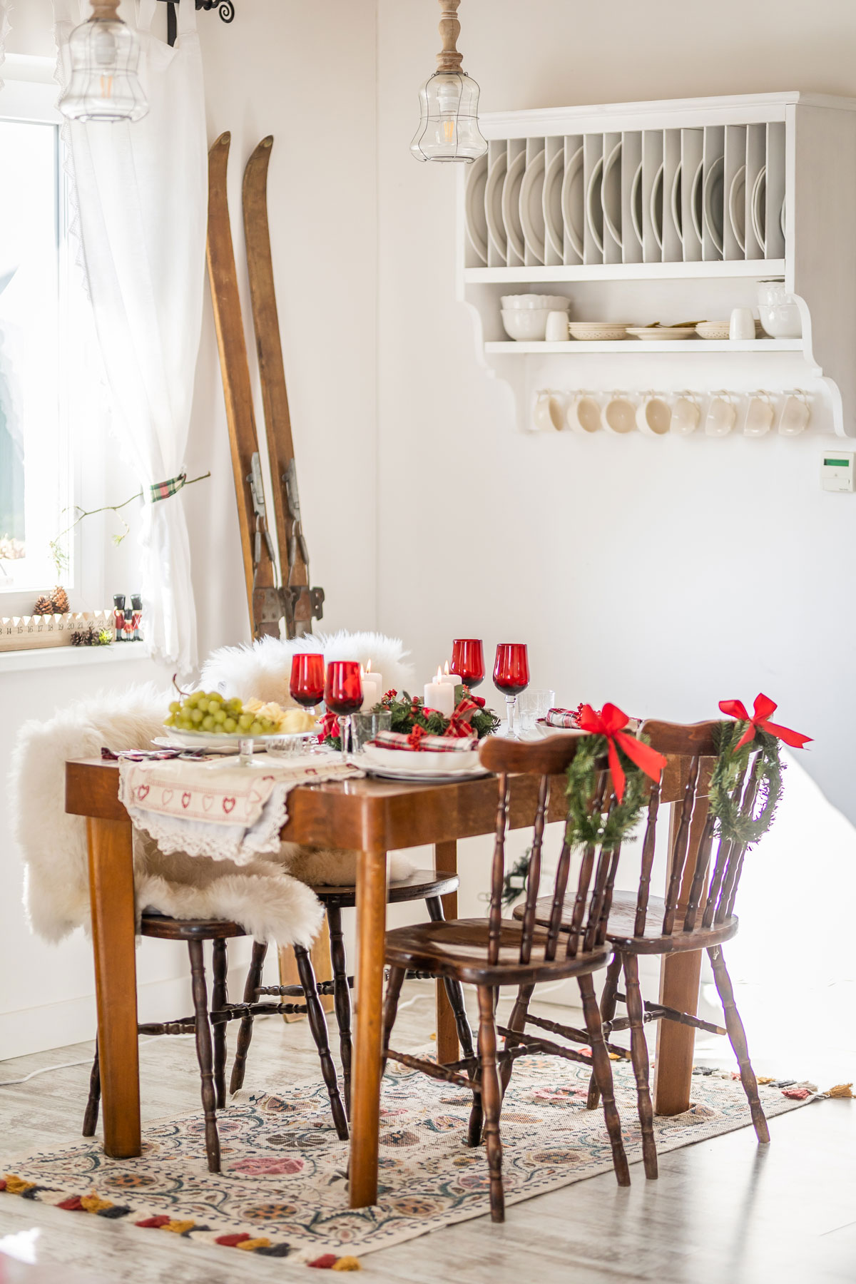 starý jedálenský stôl s retro drevenými ozdobne vyrezávanými stoličkami, na stole je slávnostné vianočné prestieranie v bielo-červeno.-zelenej kombinácii, na operadlách stoličiek sú čečinové venčeky, pri jednej stene vedľa stola sú opreté ako dekorácia retro drevené lyže