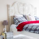 biela kovová posteľ v spálni s károvanými obliečkami, vedľa postele sú naskladané časopisy slúžiace ako stolík