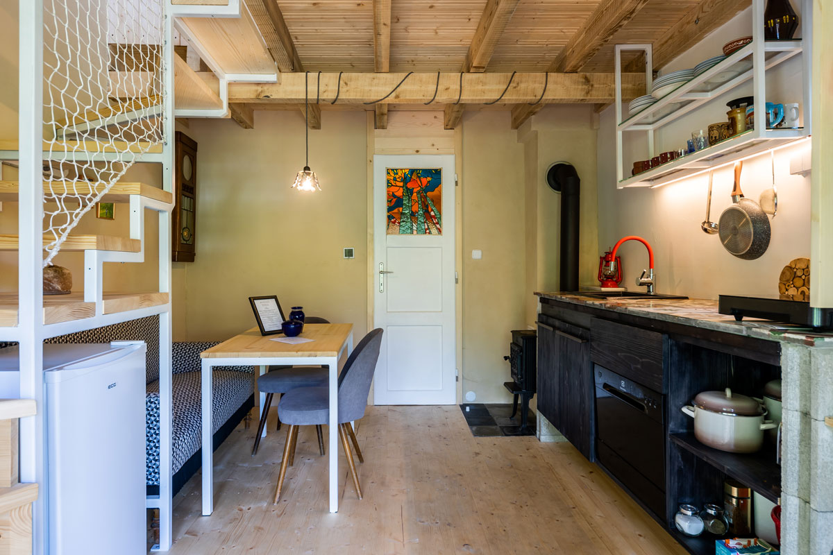 prízemie záhradnej chatky s menšou čiernou kuchynskou linkou so spotrebičmi, závesnou kovovou otvorenou policou, dreveným stropom, pod schodmi na druhé poschodie je umiestnený gauč a stôl