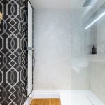 moderná kúpeľňa so vzorovanou čiernou tapetou, na stene visí moderný štíhly radiátor, oproti nemu je jednoduchý sprchový kút, podlaha je drevená