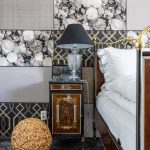 historizujúca spálňa s posteľou so zlatým kovovým čelom, vedľa nej stojí dekoratívny nočný stolík s presklenou lampou, na zemi je celoplošný čierny koberec, za posteľou je obklad tvorený z obdĺžnikov s rôznymi opakujúcimi sa vzormi