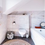 elegantná biela kúpeľňa s mramorovým obkladom so zlatým žilkovaním, kúpeľňa je doplnená o čierne akcenty v podobe zrkadla a kovového koša