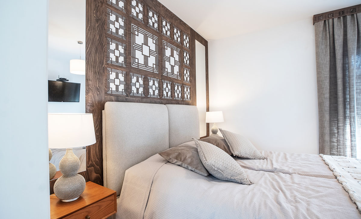 neutrálne ladená spálňa s čalúnenou posteľou, za čelom postele je drevený obklad s autorským vyrezávaným vzorom