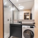 Malá kúpeľňa so sprchovacím kútom, práčkou, jednoduchým bielym obkladom, čiernou skrinkou s umývadlom a veľkým zrkadlom