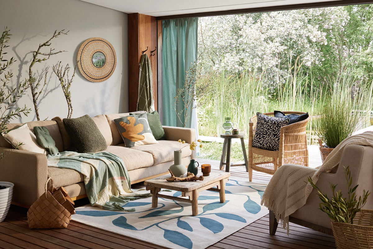 Obývačka v prírodnom boho štýle s neutrálnou sedačkou, farebnými textíliami a presklenou stenou s výhľadom do prírodnej záhrady