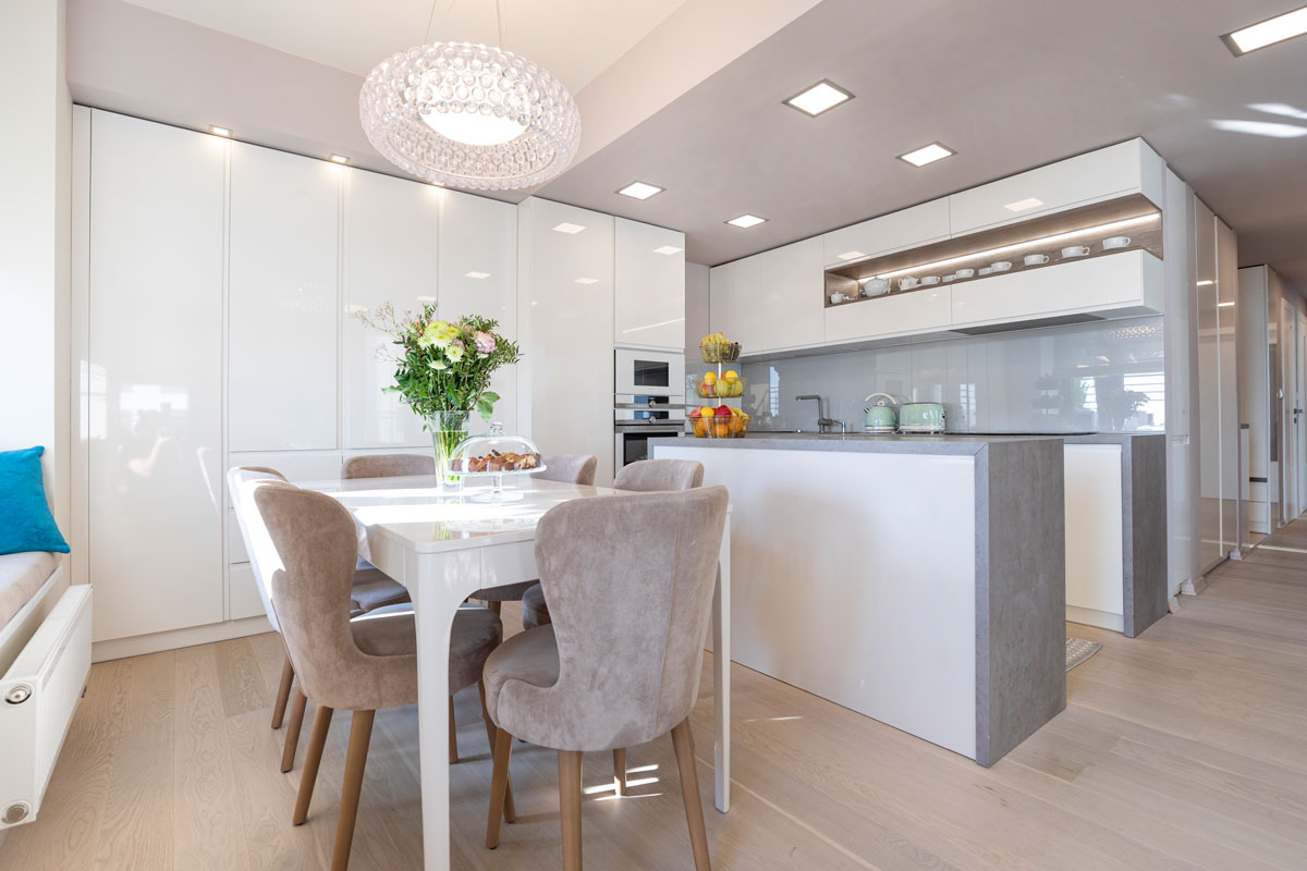 elegantná moderná kuchyňa s lesklými plochami, v bielej farbe so sivým ostrovčekom, vedľa je jedálenský stôl s čalúnenými stoličkami