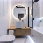 Kúpeľňa s bielym obkladom, drevenou skrinkou, umývadlom na doske a podsvieteným okrúhlym zrkadlom