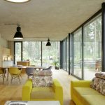 minimalistický industriálny interiér v rodinnom dome s pohľadovým betónom, so žltou sedačkou v obývačke a jedálenským stolom so žltými stoličkami, z interiéru je výhľad do prírody cez presklené steny